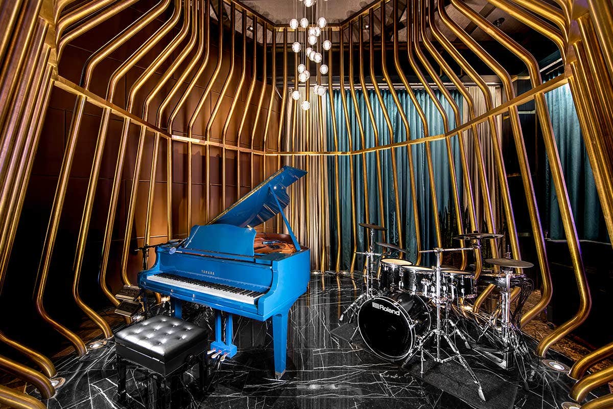 A blue grand piano and black drum set in an elegant room with golden vertical bars and a chandelier above.