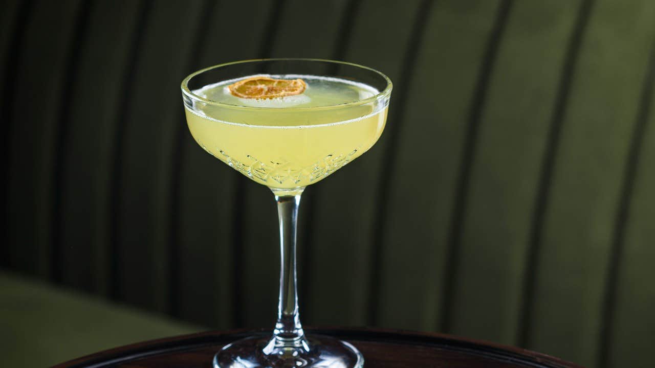 A yellow cocktail in a crystal glass garnished with a slice of dried lemon, set against a green striped background.