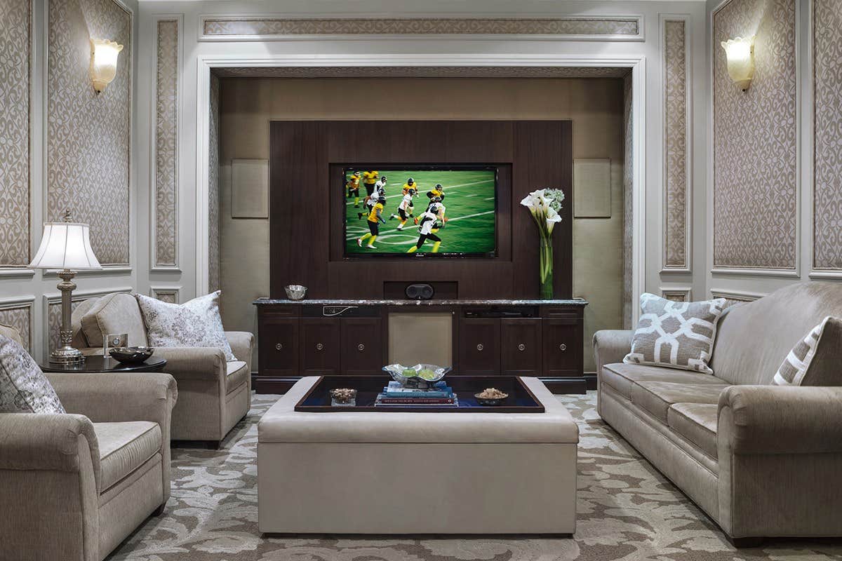 A cozy living room with two sofas, a TV showing a soccer game, a central ottoman, and a detailed patterned wallpaper.