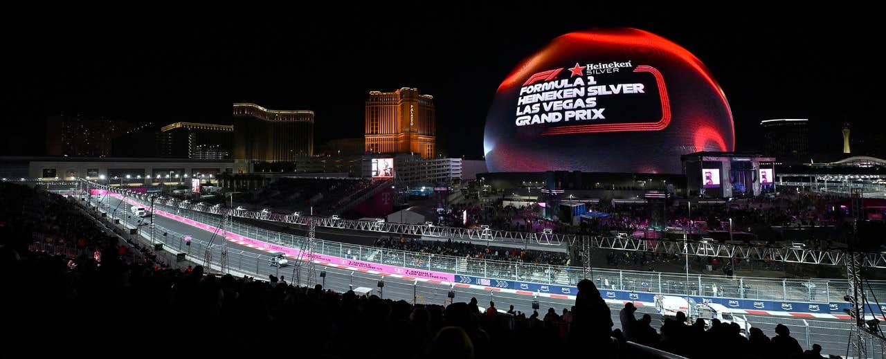 Night view of the Las Vegas Grand Prix with cars on the track and the illuminated MSG Sphere displaying event information.