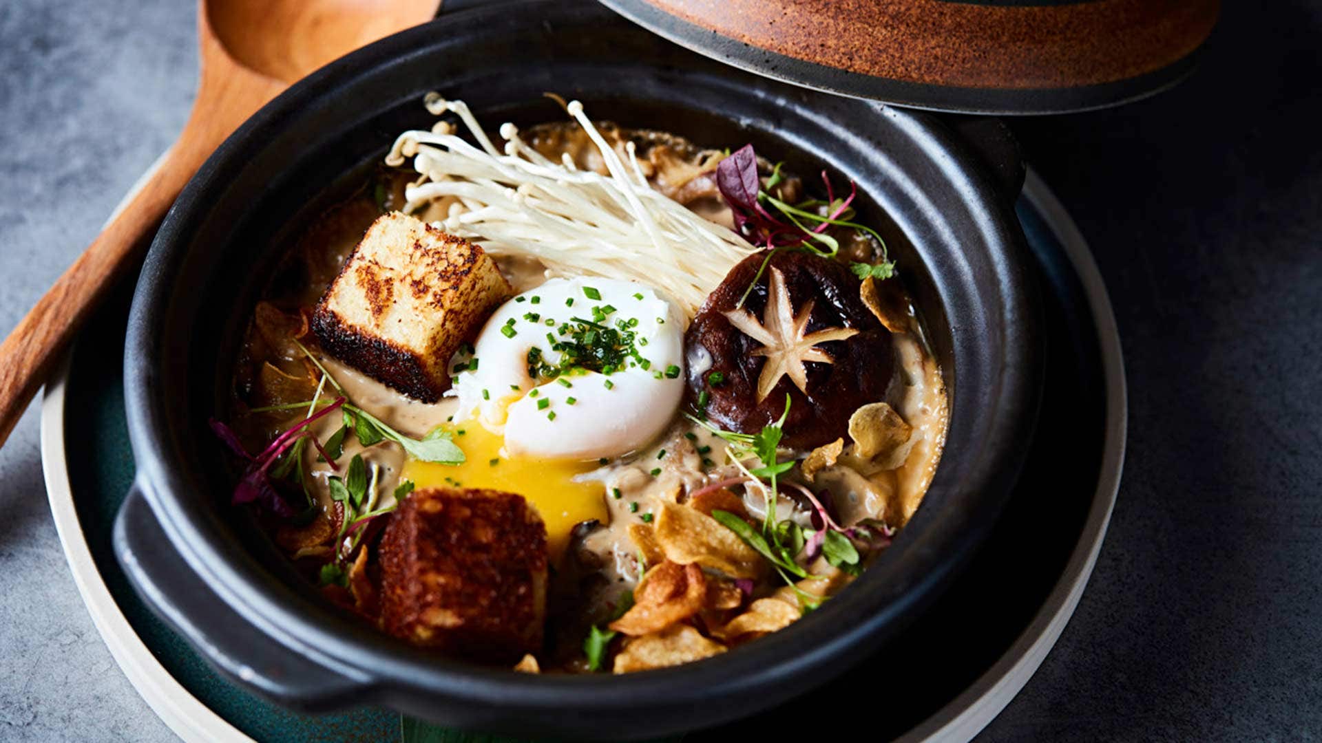 A bowl of gourmet ramen topped with soft-boiled egg, enoki mushrooms, tofu cubes, and various garnishes.