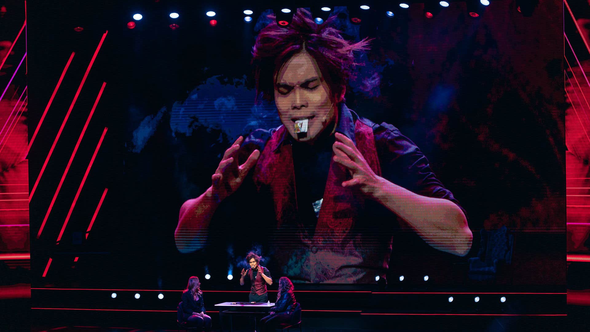 Shin Lim performing on stage with a large screen displaying his image behind him, showing his intense expression and hands raised dramatically.