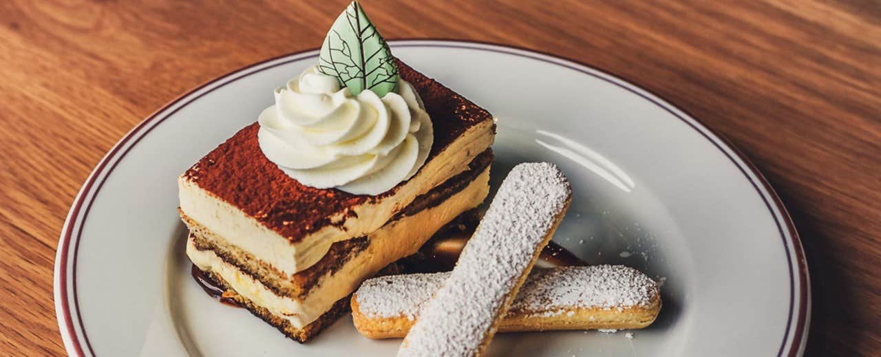 Delicious tiramisu topped with whipped cream and a decorative leaf, served with two powdered sugar-covered ladyfingers.