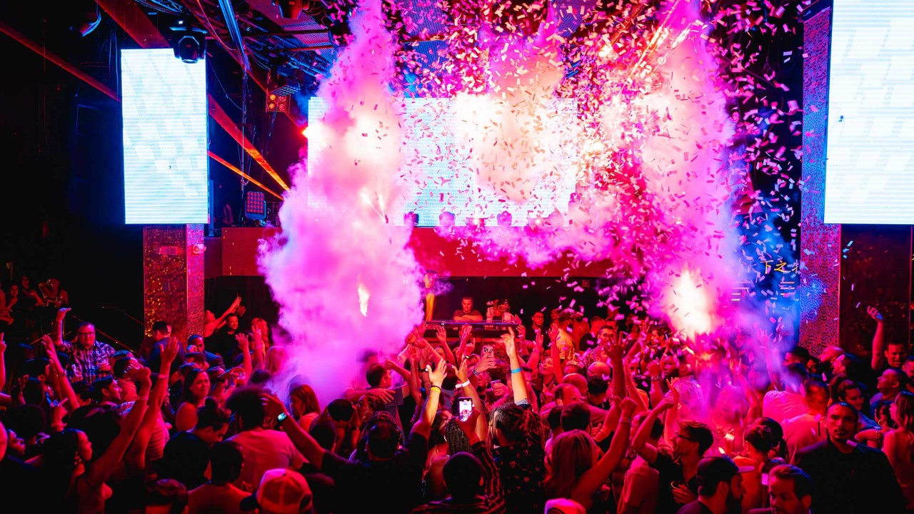 Crowded nightclub with colorful lights, confetti, and smoke effects as people dance and cheer near a DJ booth.