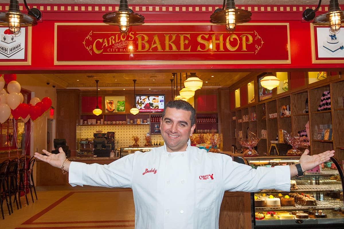 A man in a white chef's coat stands with open arms in front of a bakery named "Cake Boss Bake Shop.