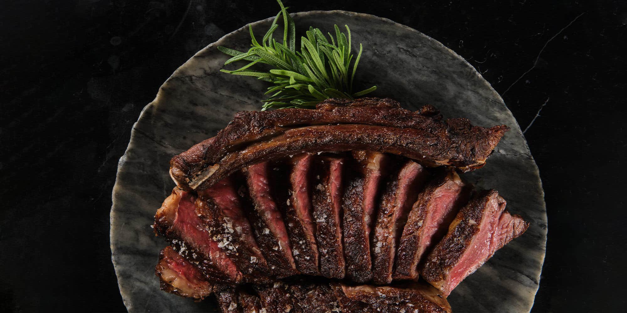 Ribeye sliced and arranged along with herbs on a dark gray plate, laying on a dark surface