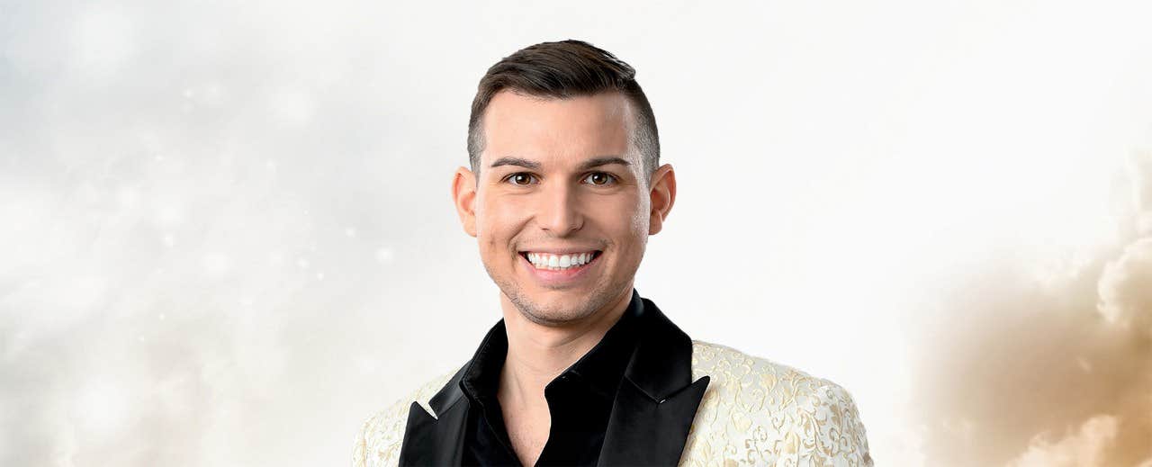 A man in a patterned white suit jacket and black shirt smiles against a light cloudy background.