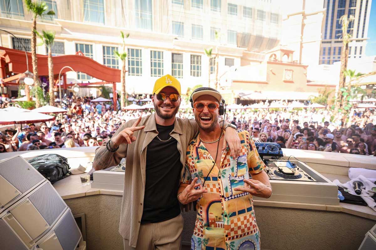 Two men smiling and posing in front of a cheering crowd near DJ equipment at an outdoor event.