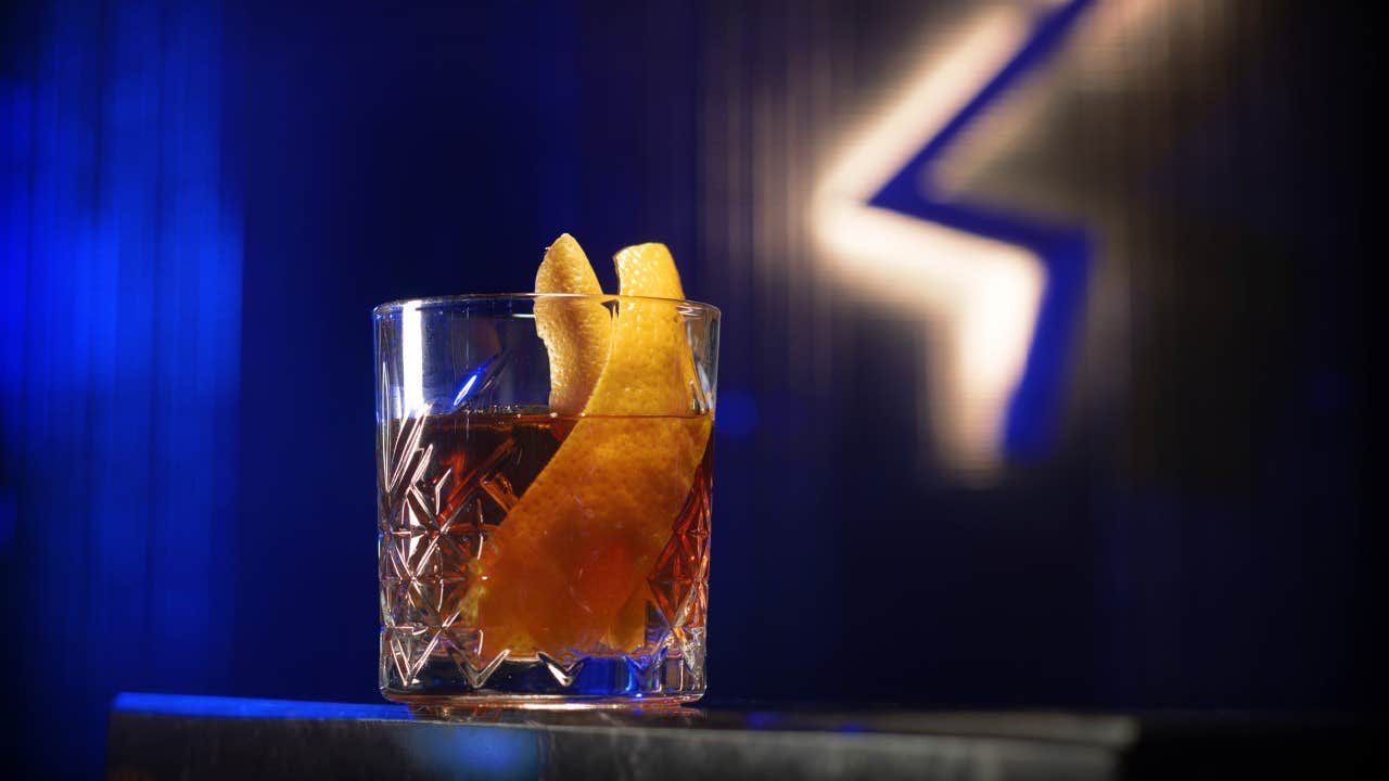 A glass of amber-colored cocktail garnished with orange peel, with a blue and white neon light in the background.