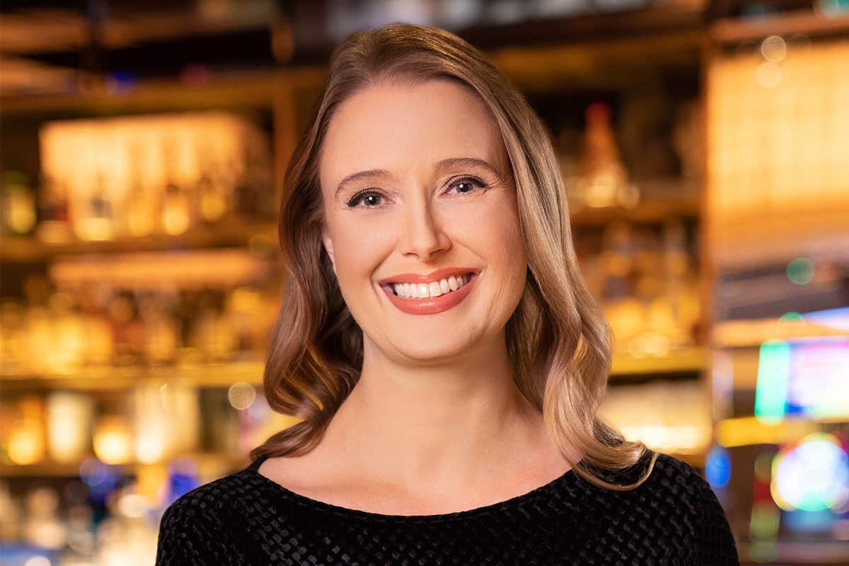 A smiling woman with long hair stands in front of a warmly lit background, wearing a black top.