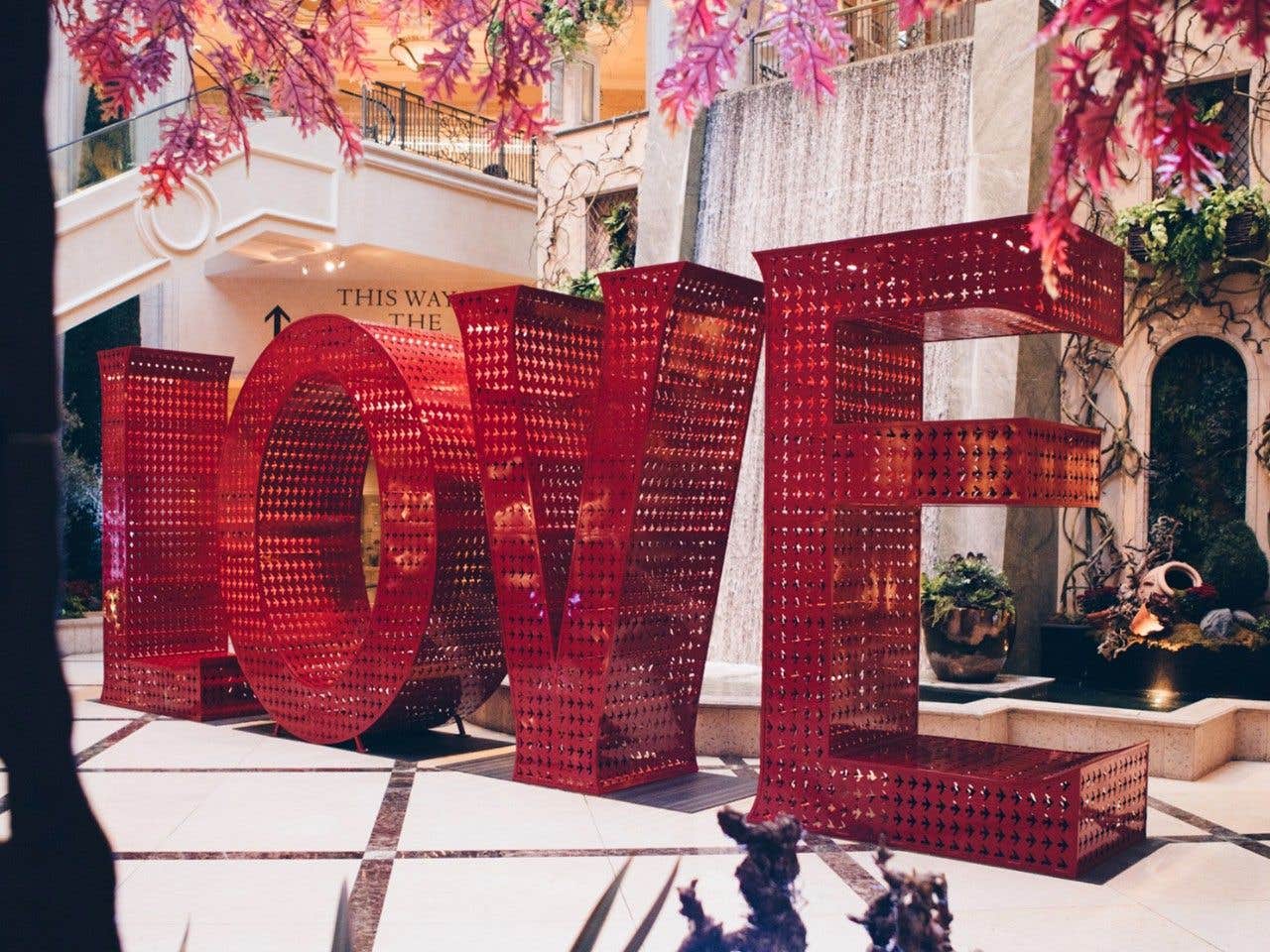 Large red "LOVE" sculpture in an indoor garden setting with a small waterfall and decorative plants around it.