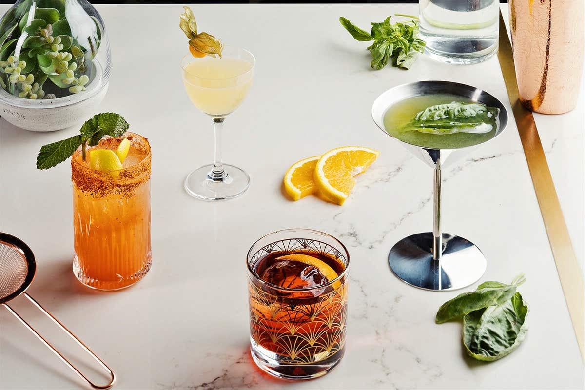 A variety of colorful cocktails on a marble table, garnished with herbs, citrus slices, and a plant in the background.