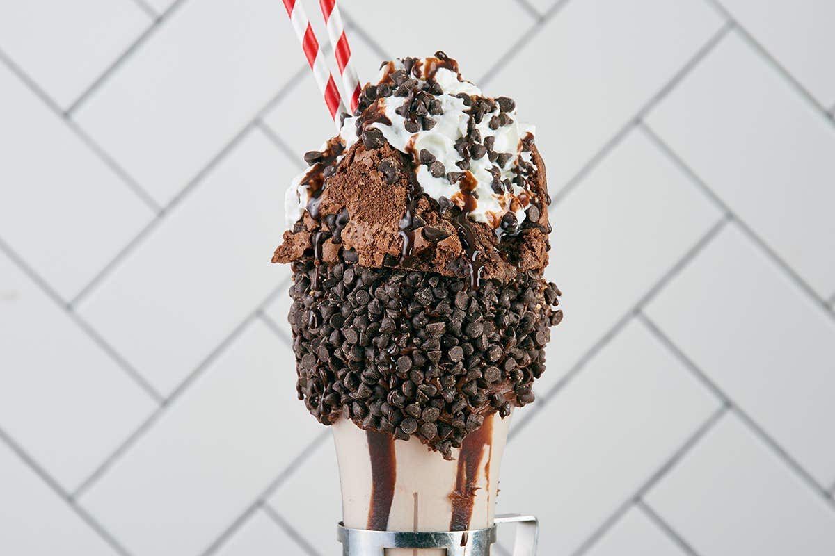 Chocolate milkshake topped with whipped cream, chocolate drizzle, brownies, and mini chocolate chips, with striped straws.