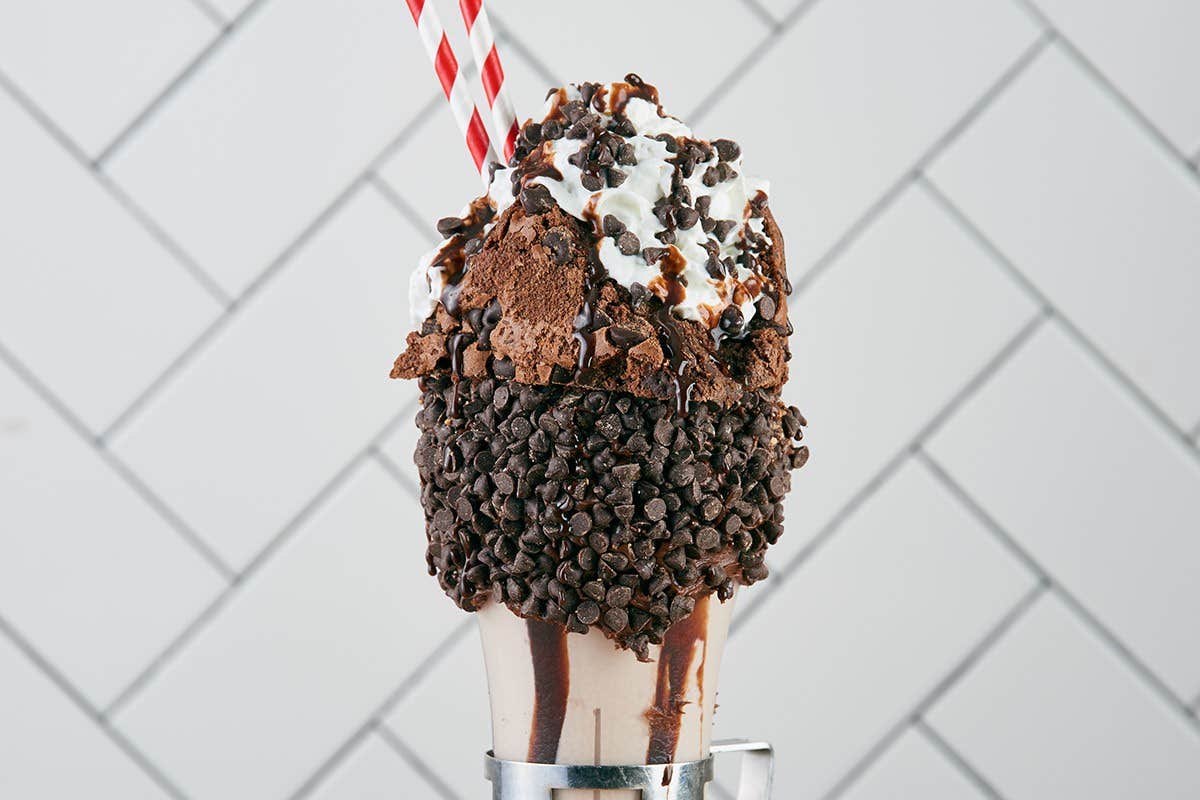 Chocolate milkshake topped with whipped cream, chocolate drizzle, brownies, and mini chocolate chips, with striped straws.