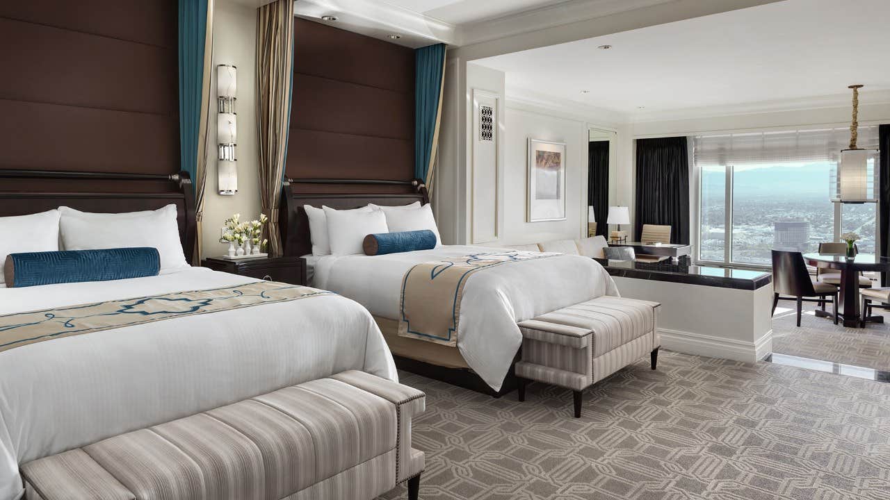 Luxurious hotel room with two large beds, a seating area, elegant decor, large windows, and a city view.