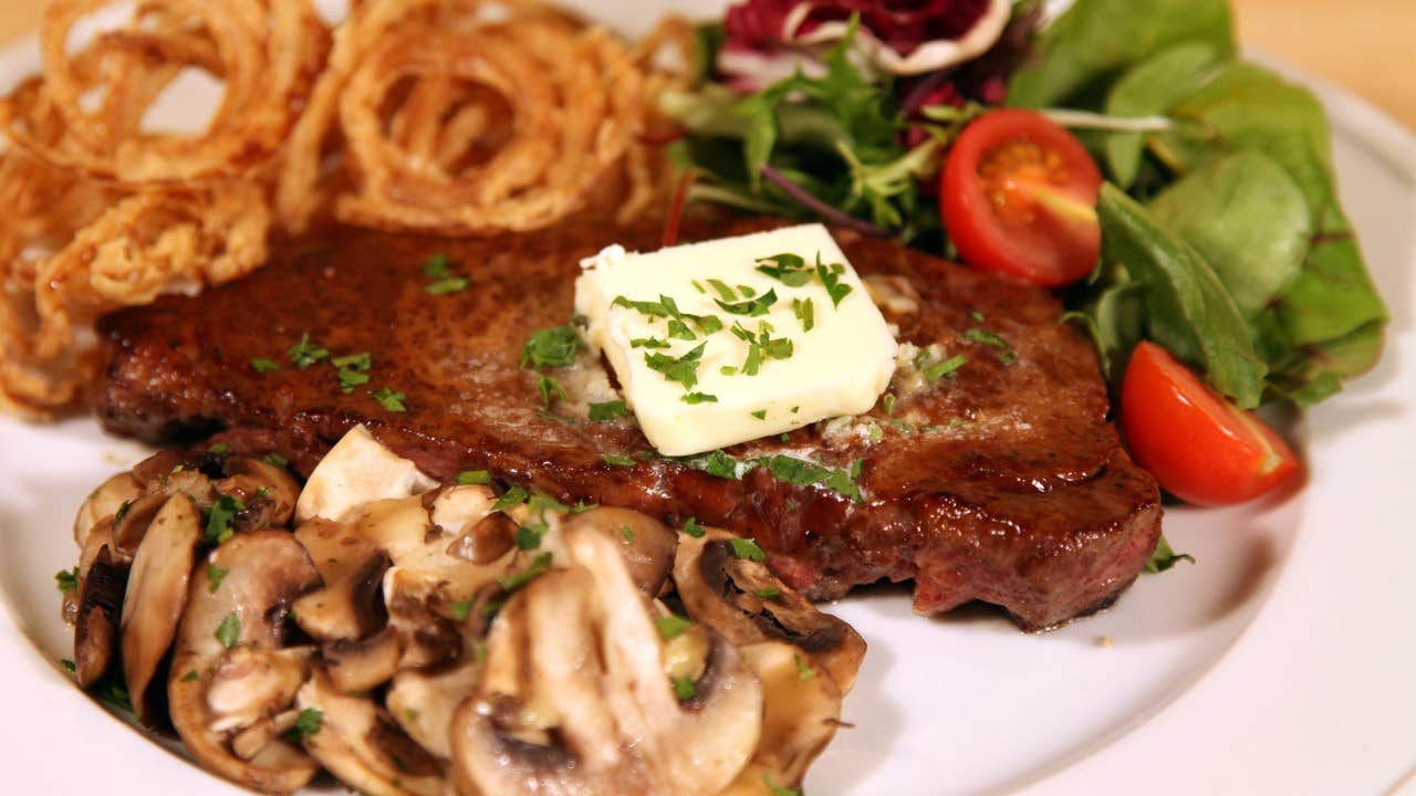 A plate with a steak topped with butter, mushrooms, onion rings, and a fresh salad with cherry tomatoes.