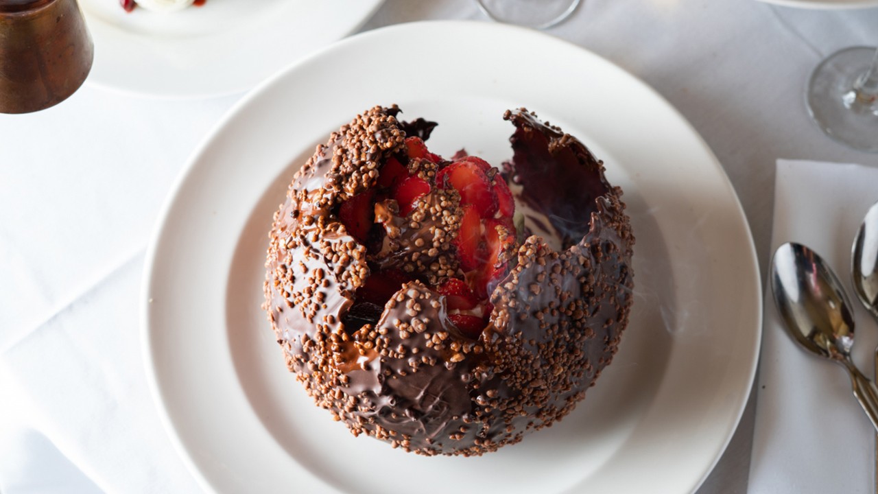 A cracked chocolate sphere revealing strawberries, served on a white plate with spoons and napkins beside it.