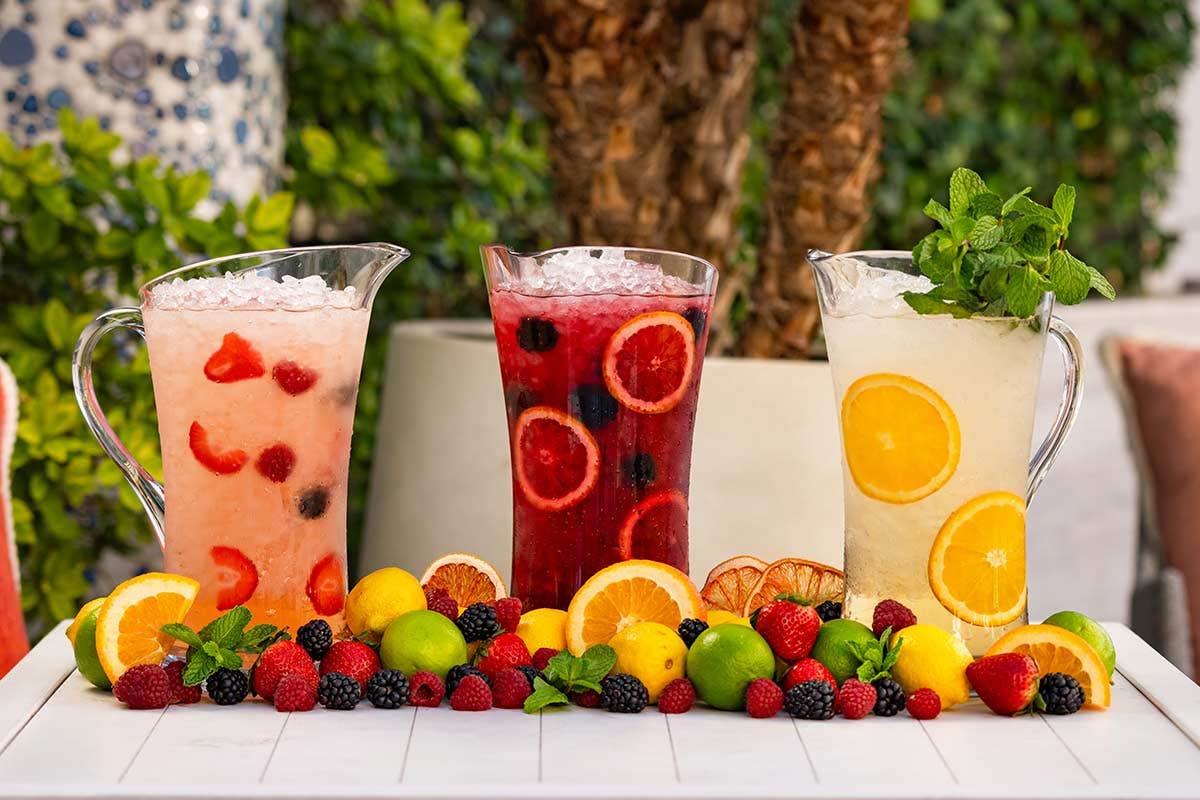 Three pitchers of infused water with assorted fresh fruits on a white table, surrounded by various colorful fruits.