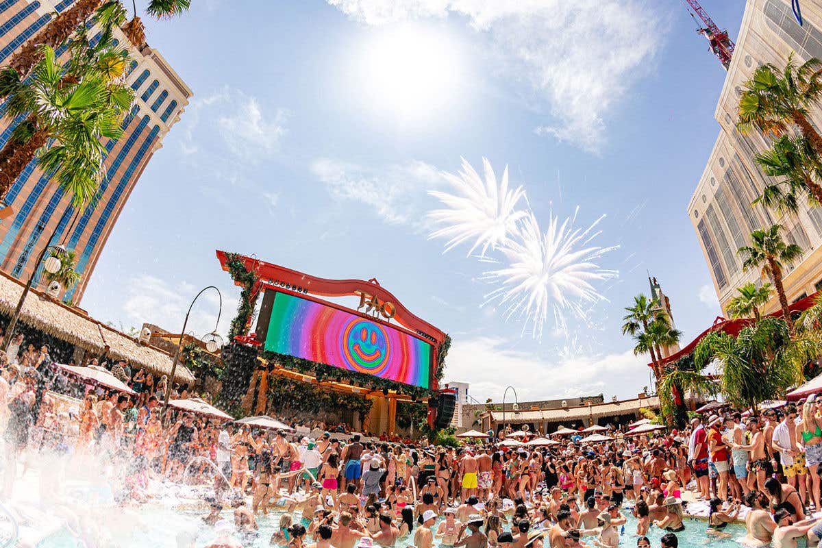 A crowded pool party at TAO Beach under a sunny sky with fireworks, featuring a large LED screen showing a rainbow smiley face.