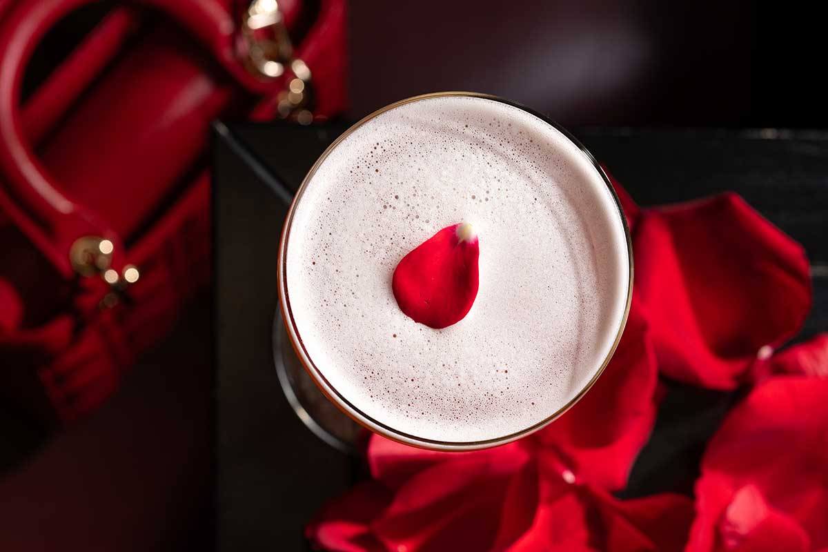 Frothy drink with a red rose petal on top and scattered red rose petals around it.