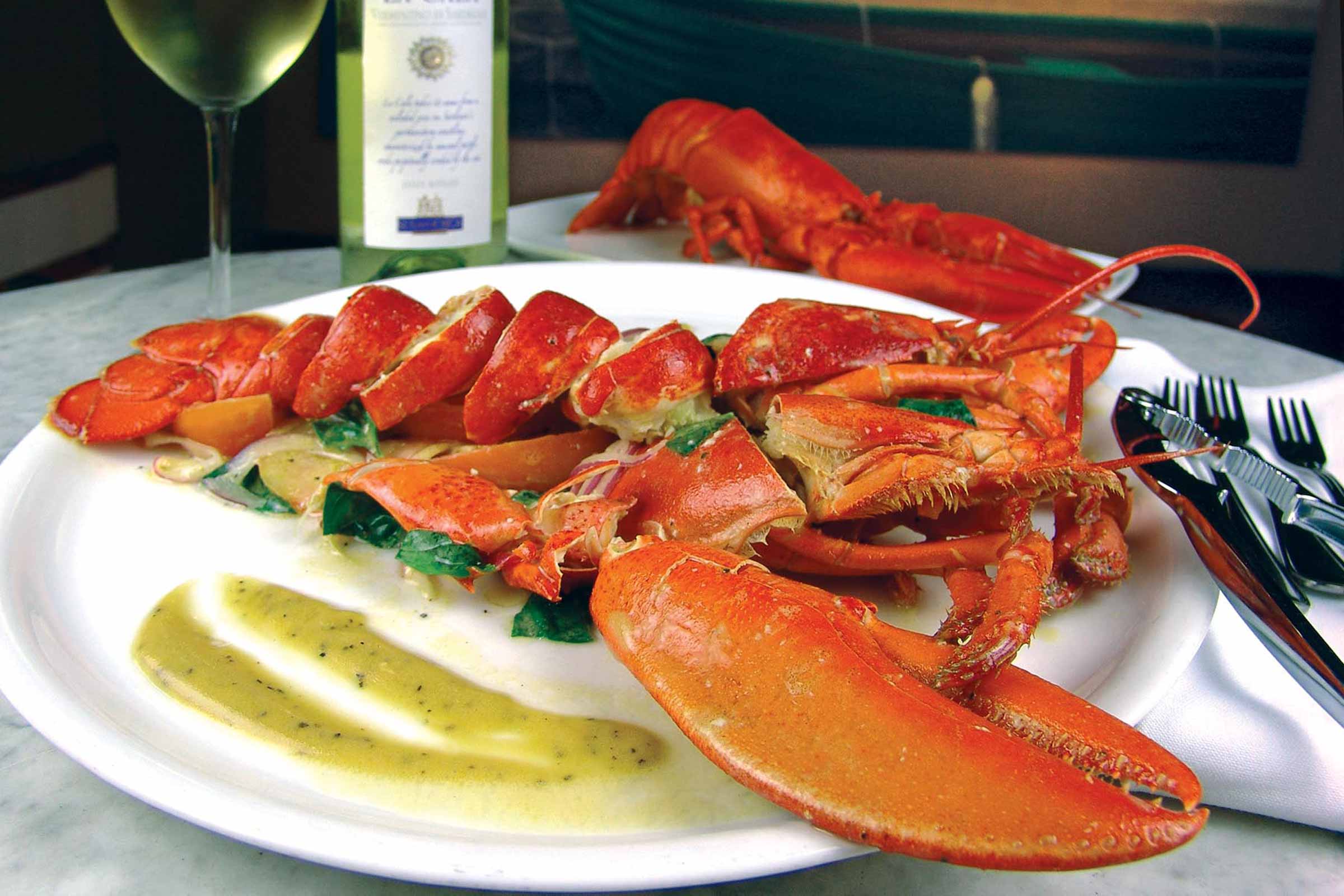 A plated lobster dish with garnish and sauce, accompanied by utensils and a glass and bottle of white wine.