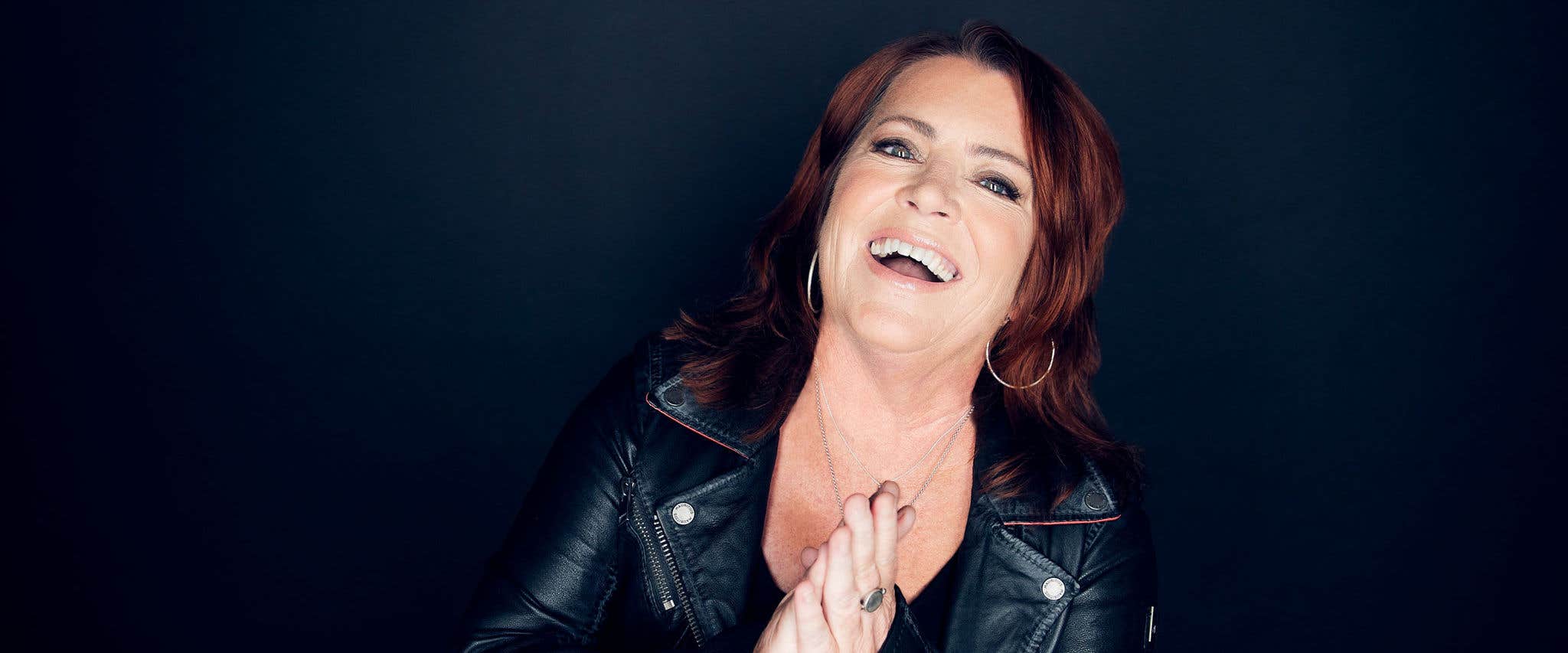 Kathleen Madigan with long hair, wearing a leather jacket, smiling and clapping her hands against a dark background.