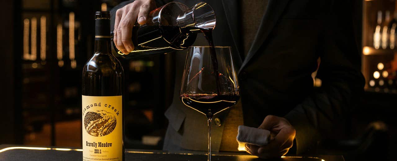 Person pouring red wine from a decanter into a glass next to a bottle of Diamond Creek 2011.