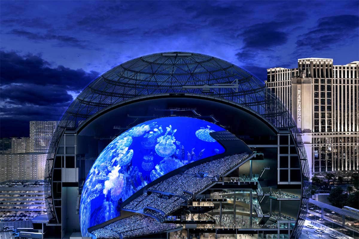 Dome-shaped concert hall at night, with a large digital screen displaying vibrant visuals and a crowd seated inside.