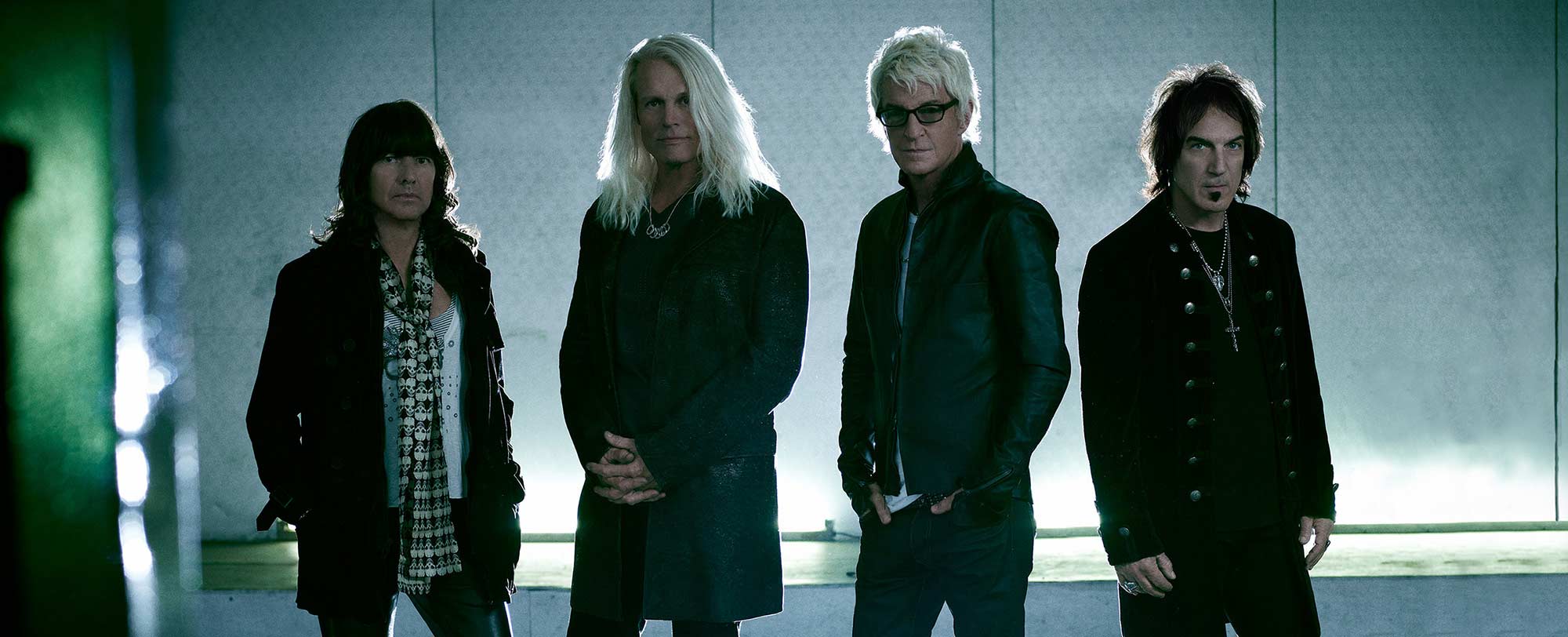 Four men standing in a row, wearing dark clothing, with a light background. The man second from left has long white hair.