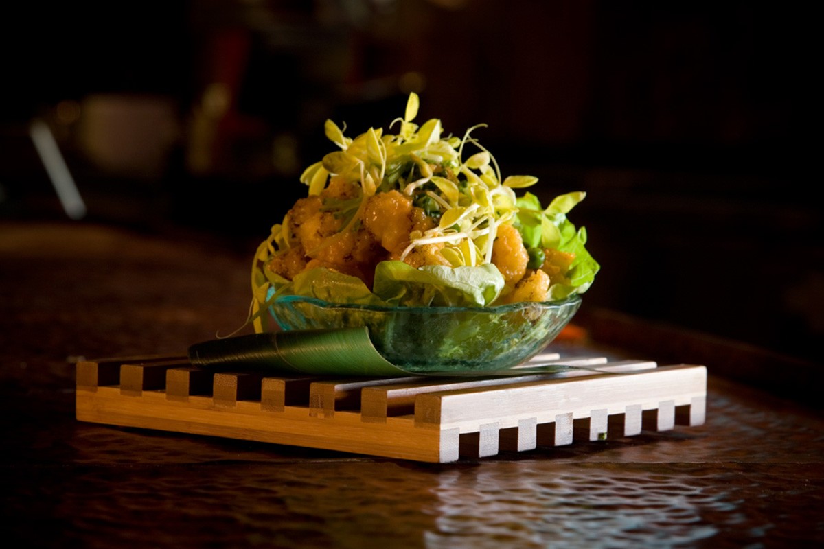 A glass bowl of vegetable salad garnished with pea shoots, placed on a wooden tray on a table.