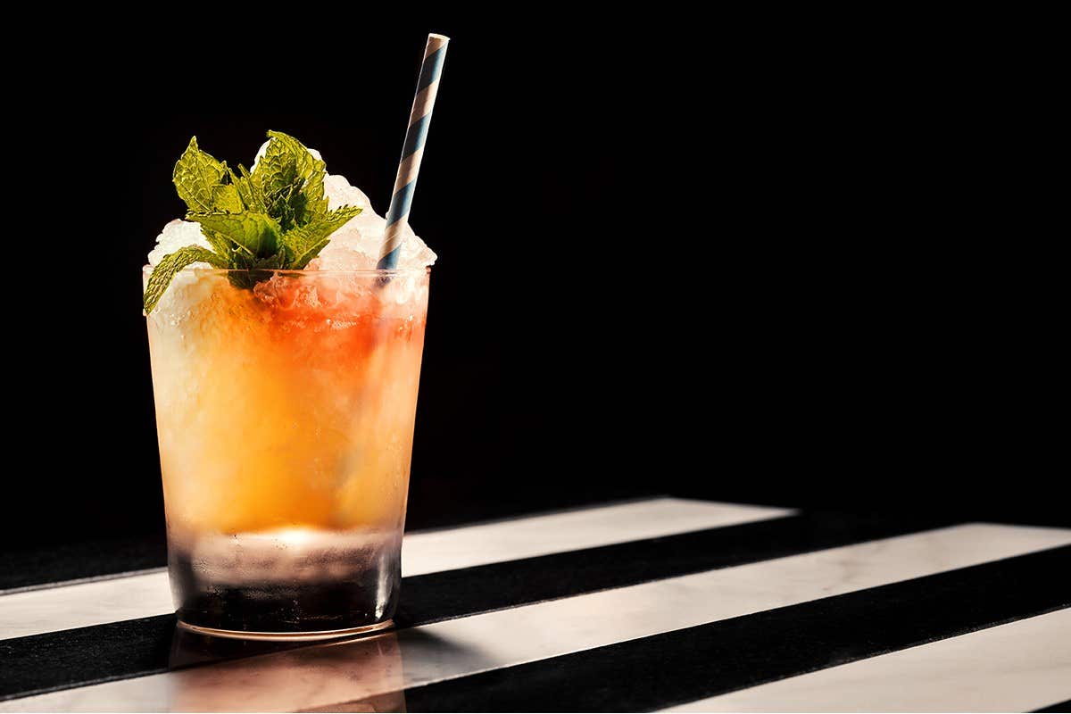 A cocktail in a glass with a straw, garnished with mint leaves and crushed ice, on a black and white striped surface.