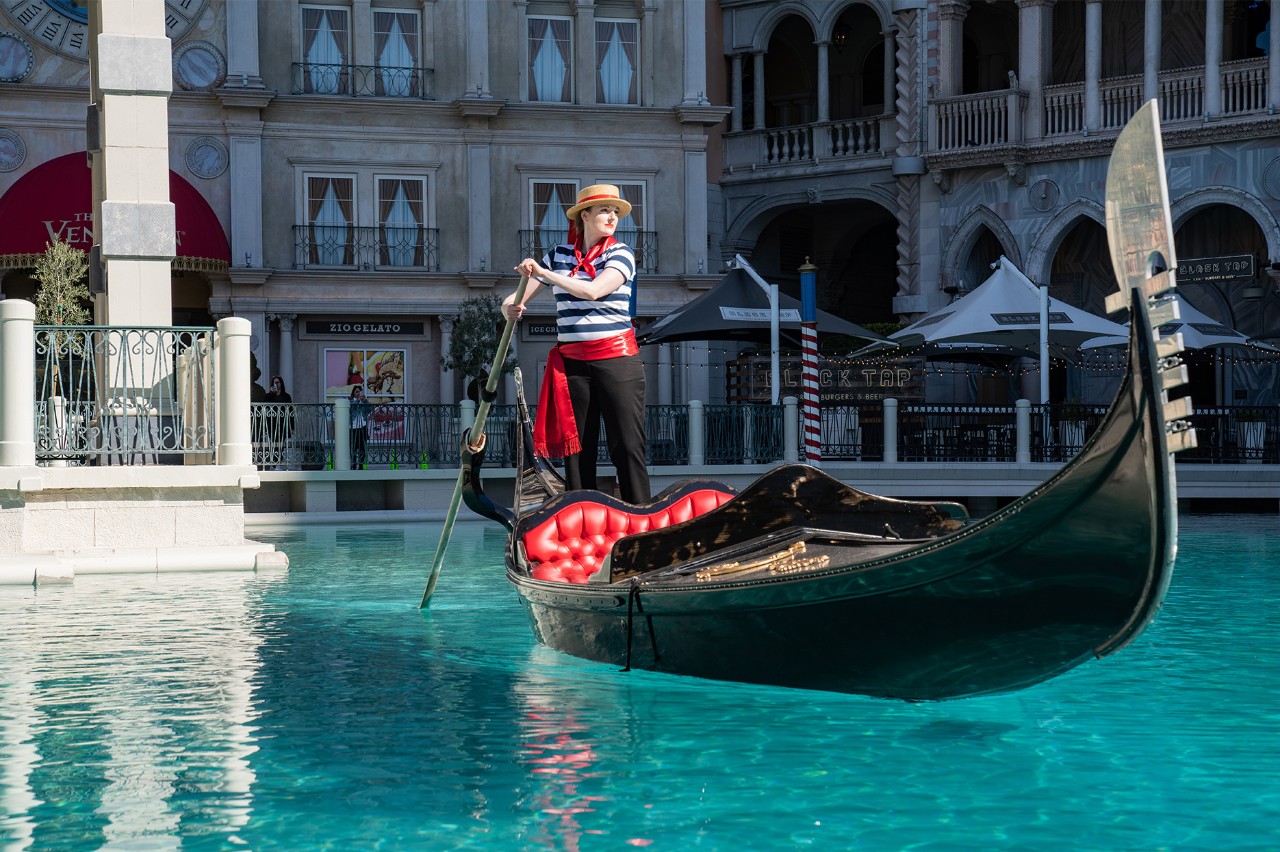 A gondolier in traditional attire stands on a gondola in a turquoise canal, with Venetian-style buildings in the background.