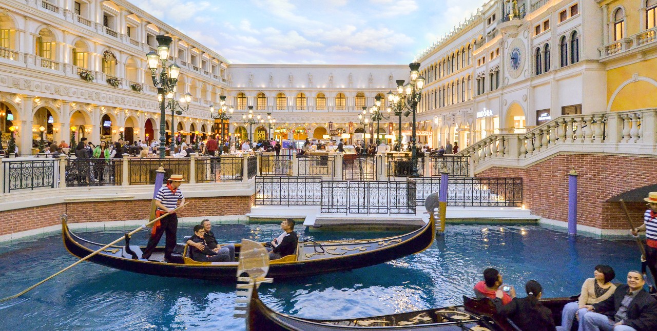 Indoor canal with gondolas, passengers, and a gondolier. European-style architecture and crowds in the background.