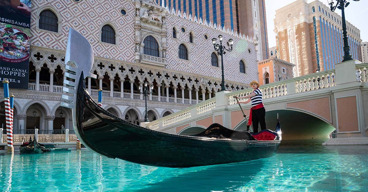 A gondolier rows a black gondola on a clear turquoise canal in front of a Venetian-style building.