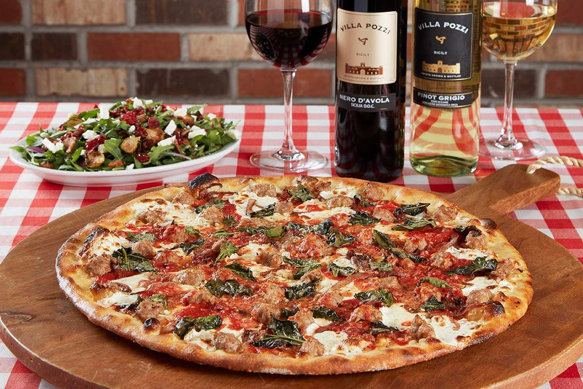 A freshly baked pizza on a wooden board, a salad, two bottles of wine, and two wine glasses on a checkered tablecloth.