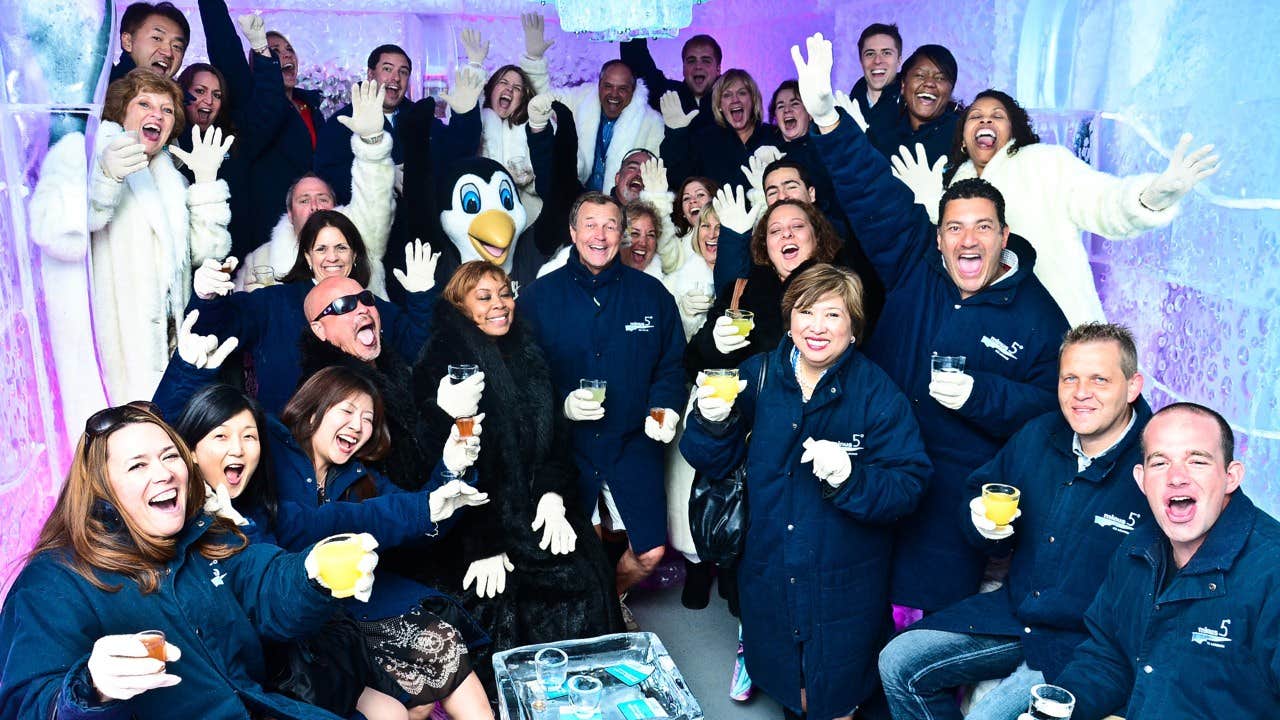 A large group of people, dressed in winter coats, cheerfully posing in an ice bar with colorful lighting.