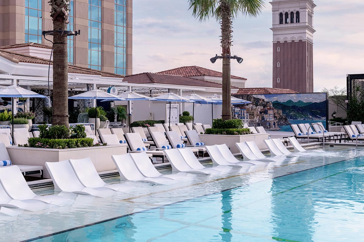 A luxurious poolside with white sun loungers, umbrellas, palm trees, and a tall tower in the background.