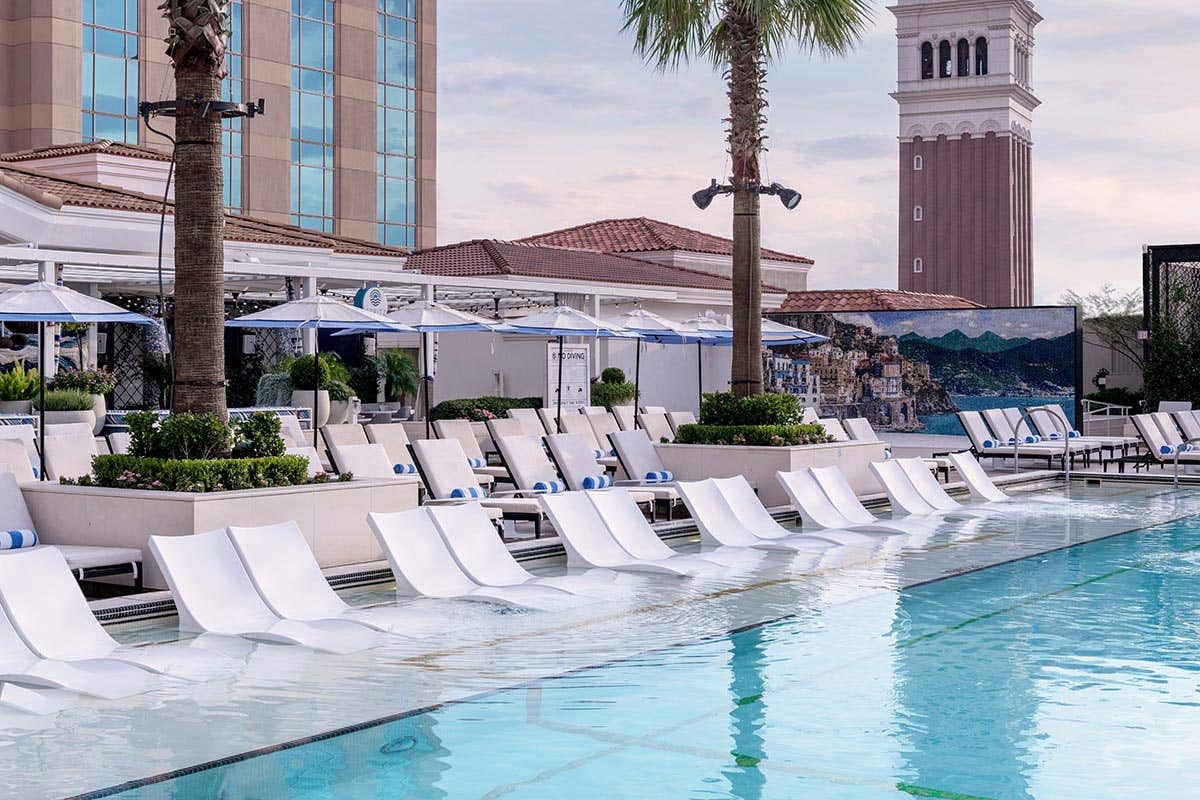 A luxurious poolside with white sun loungers, umbrellas, palm trees, and a tall tower in the background.