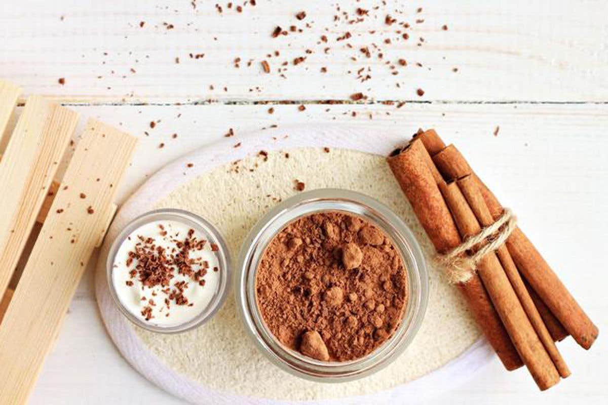 Glass jar of cocoa powder with a bowl of cream and cinnamon sticks on a wooden surface.