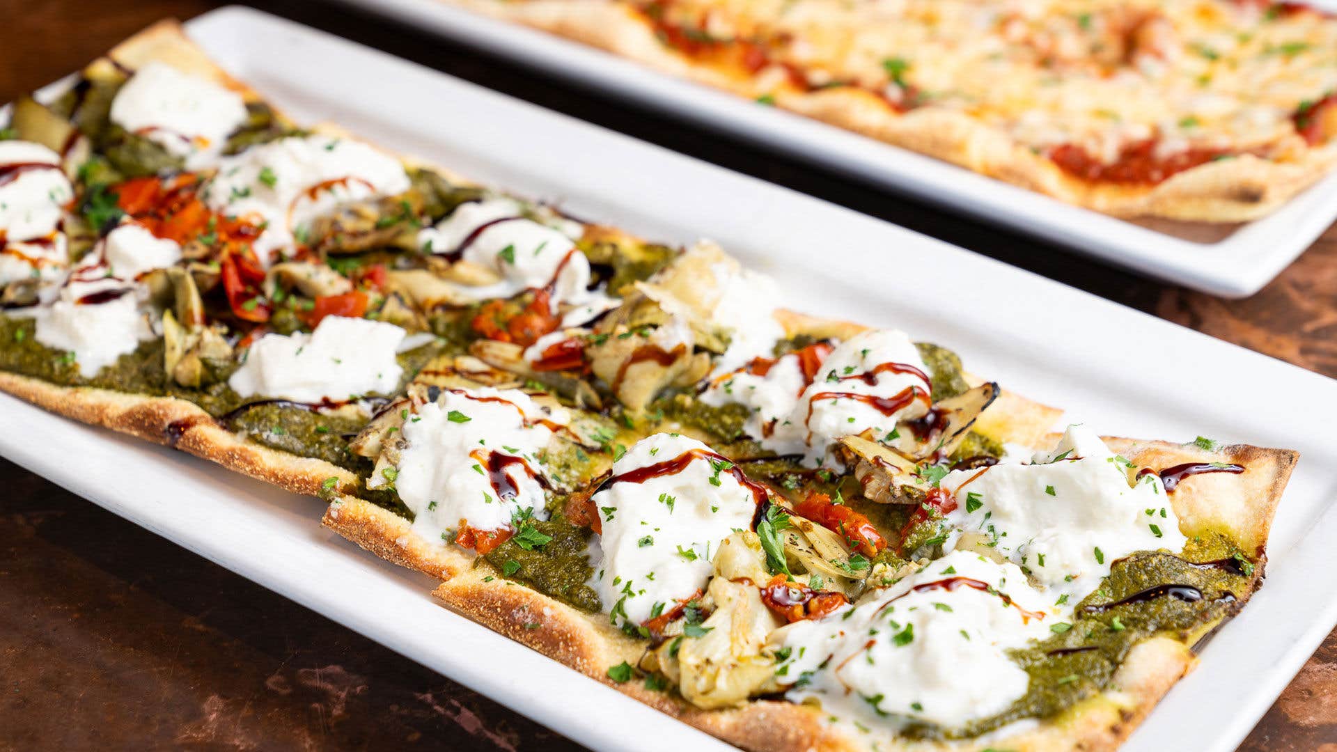 Flatbread pizza with pesto, cheese, artichokes, roasted tomatoes, and balsamic glaze on a white plate.