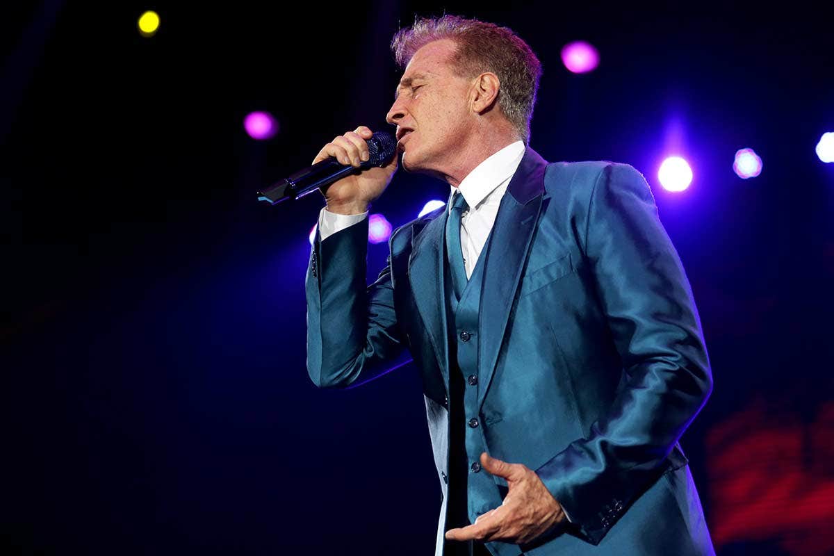 A man in a blue suit passionately singing into a microphone on a lit stage.