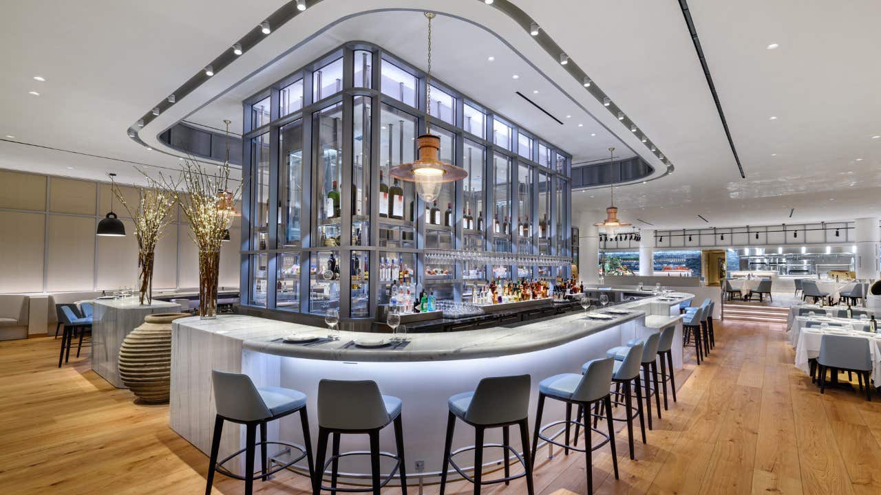 Modern restaurant with central bar, high stools, large windows, and elegant decor, featuring light wood floors and white walls.