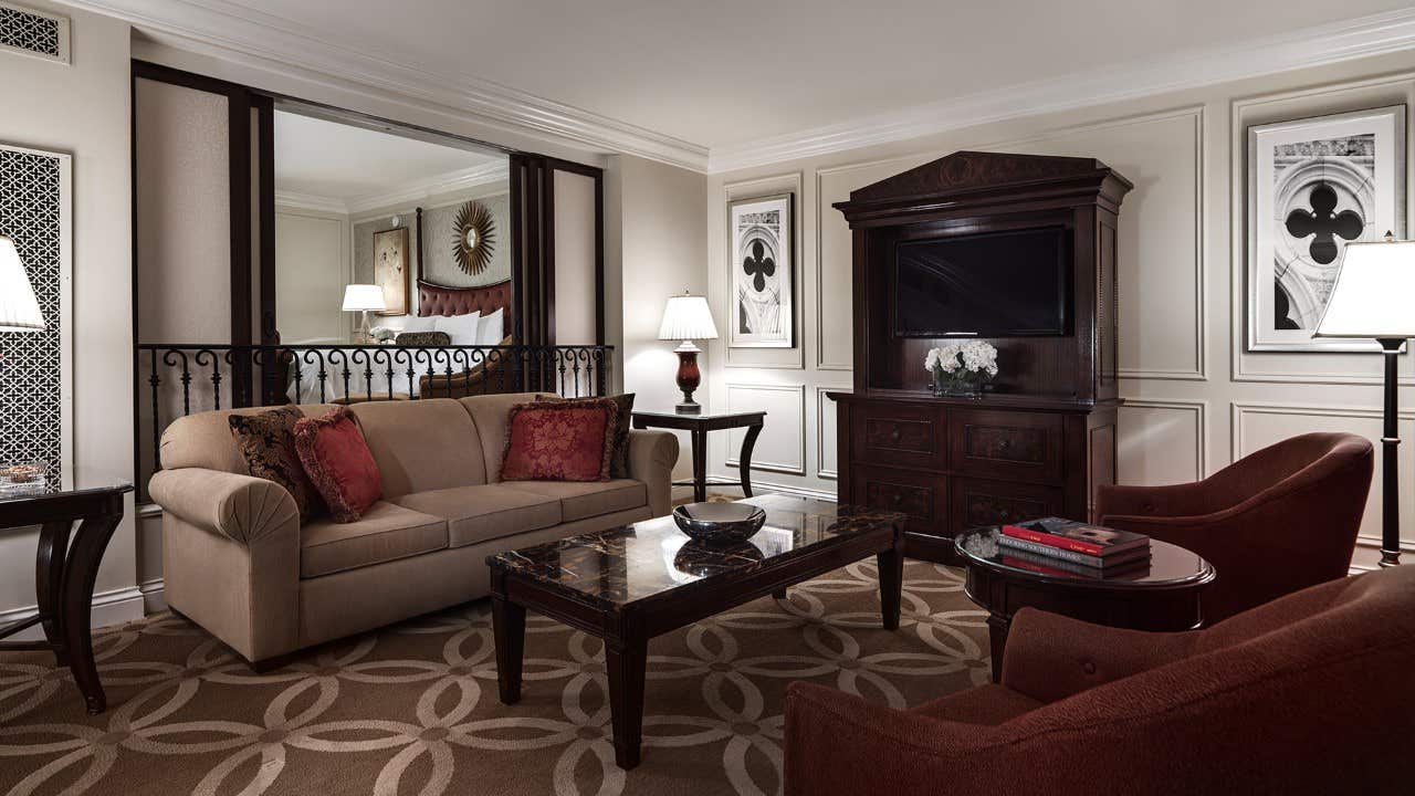 Elegant hotel room with a beige sofa, red armchair, coffee table, and TV console, featuring classic decor accents.