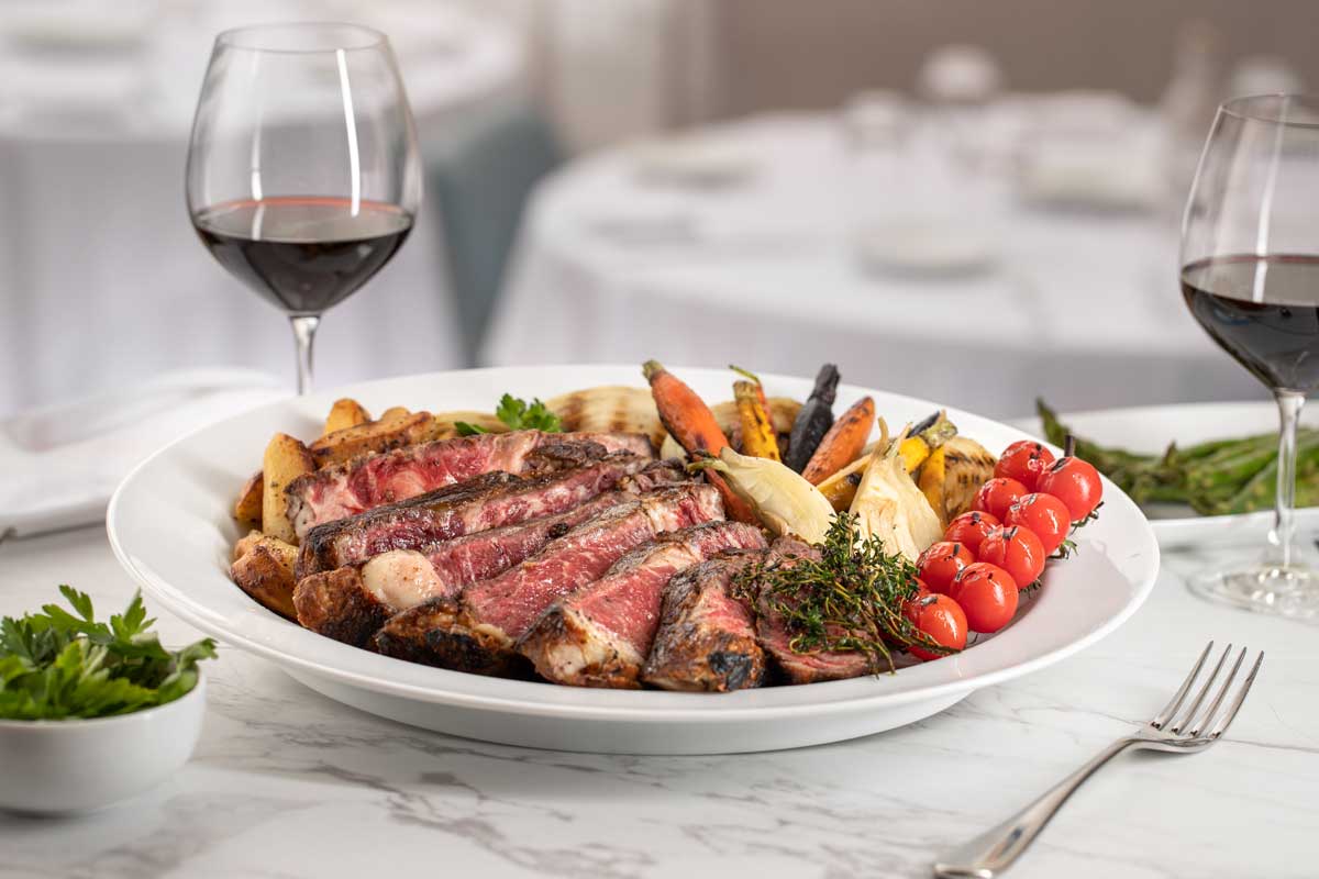 A plate of grilled meat and vegetables is served alongside two glasses of red wine on a marble table.