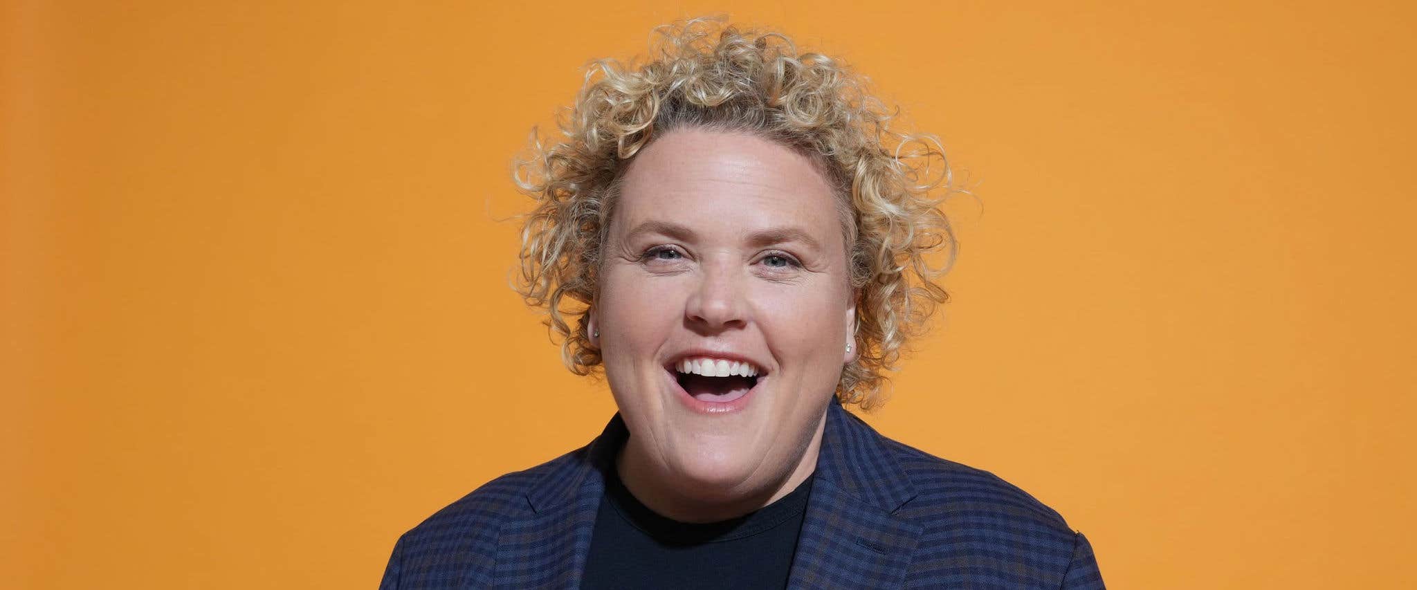 Fortune Feimster smiling, wearing a dark blue plaid blazer and black shirt, set against an orange background.