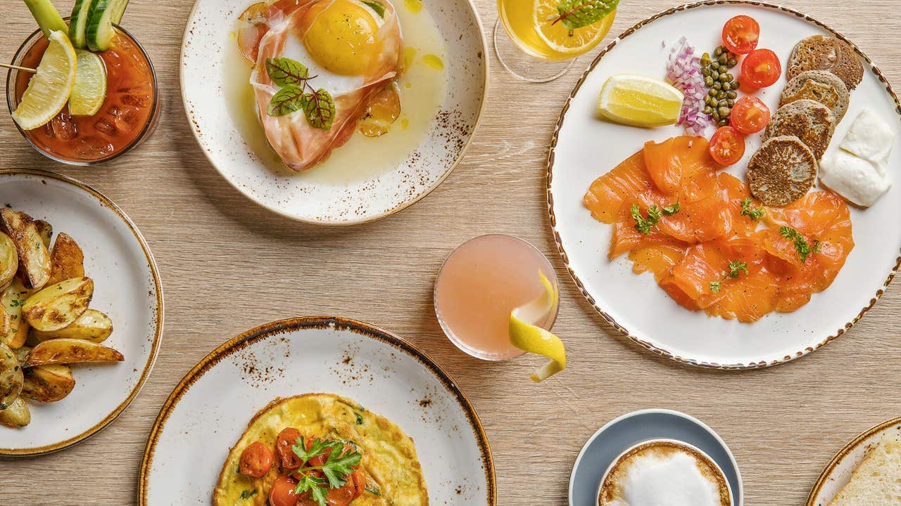 Plates of assorted gourmet dishes with smoked salmon, poached egg, cocktail, latte, and seasoned potato wedges on a table.