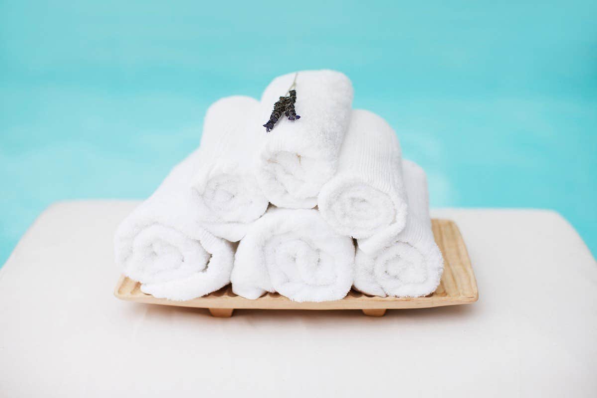 A stack of six rolled white towels with a sprig of lavender on top, placed on a wooden tray against a blue background.