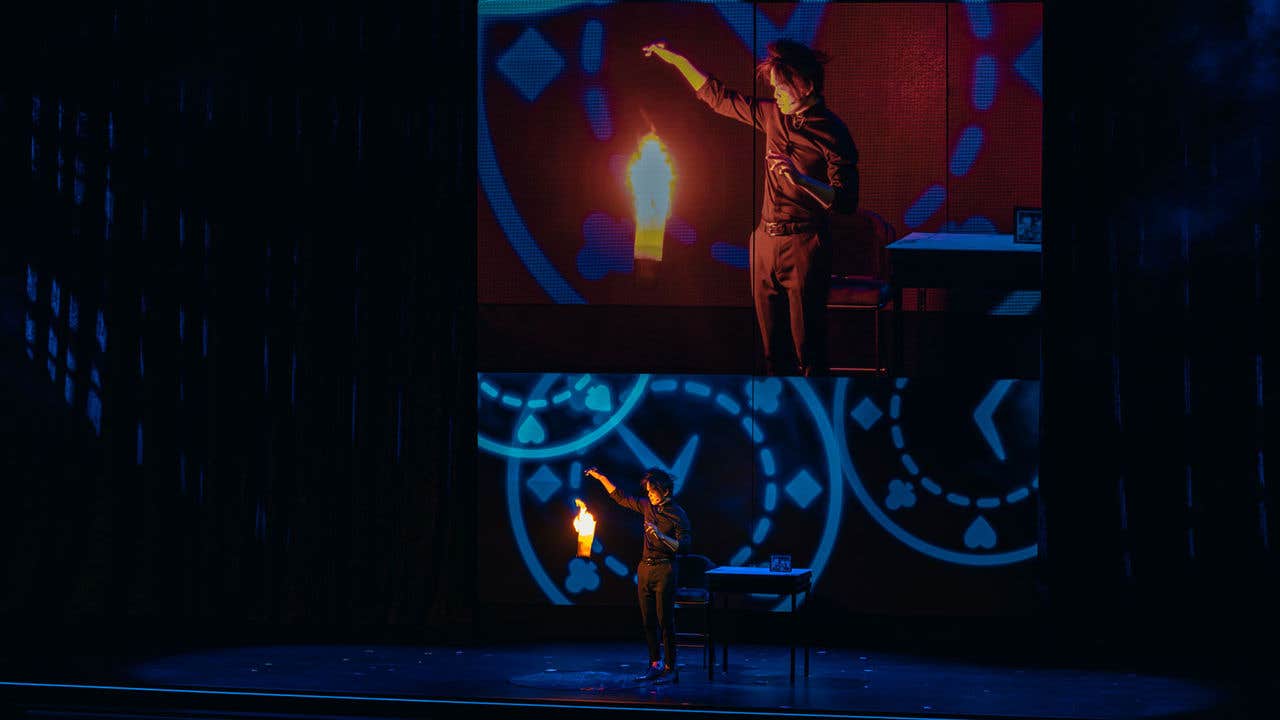 Shin Lim on stage with a fiery torch, projected on a large screen with blue abstract patterns.