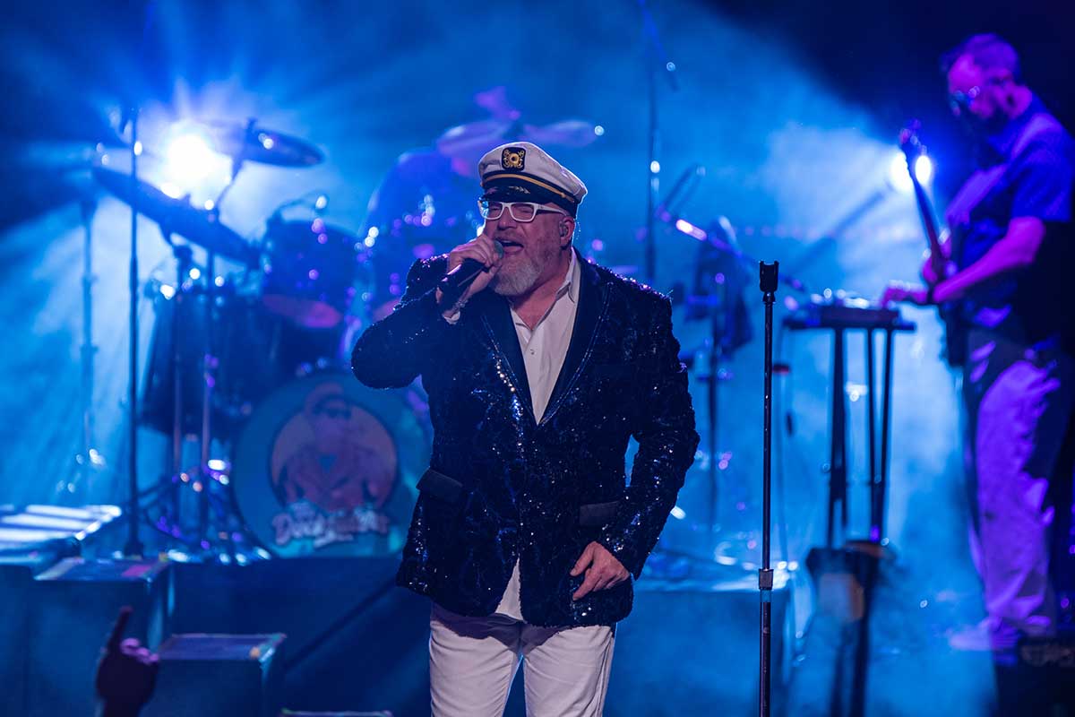 Singer performing on stage wearing a captain's hat and sequined jacket, with band members and bright lights in the background.