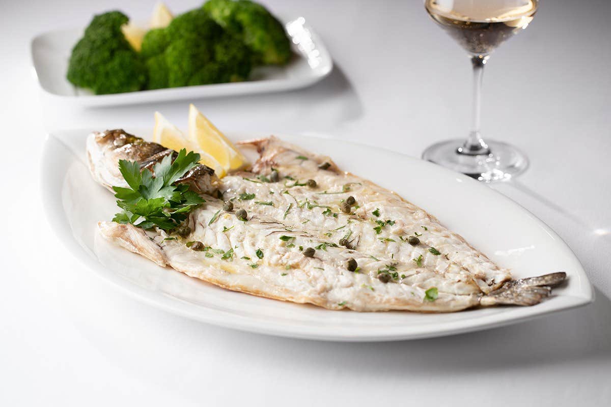 Grilled fish garnished with parsley and capers on a white plate, with lemon wedges, broccoli, and a glass of white wine.